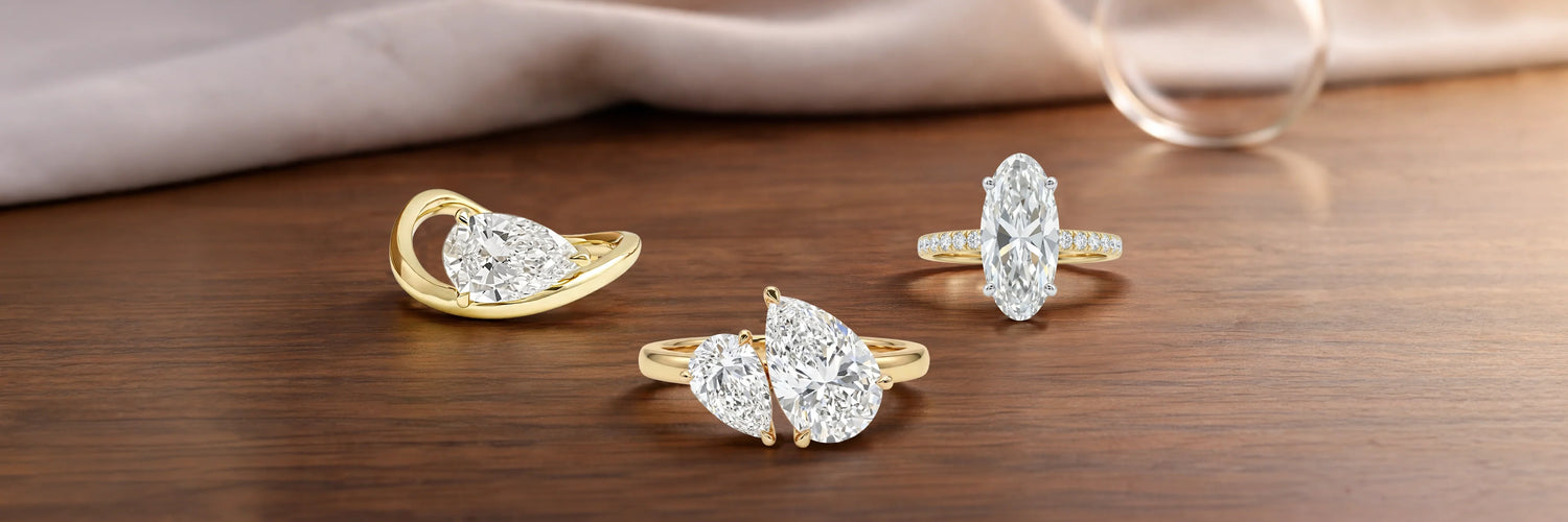 Three unique lab-grown diamond engagement rings in yellow gold — pear cut bypass solitaire, double pear Toi et Moi ring, and elongated oval solitaire with pavé band on dark walnut wood surface