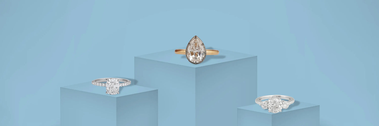 Three old cut lab-grown diamond engagement rings in yellow gold — fancy yellow oval solitaire, large old mine cushion cut solitaire with claw prongs, and fancy yellow three-stone ring with round white diamond side stones on champagne satin