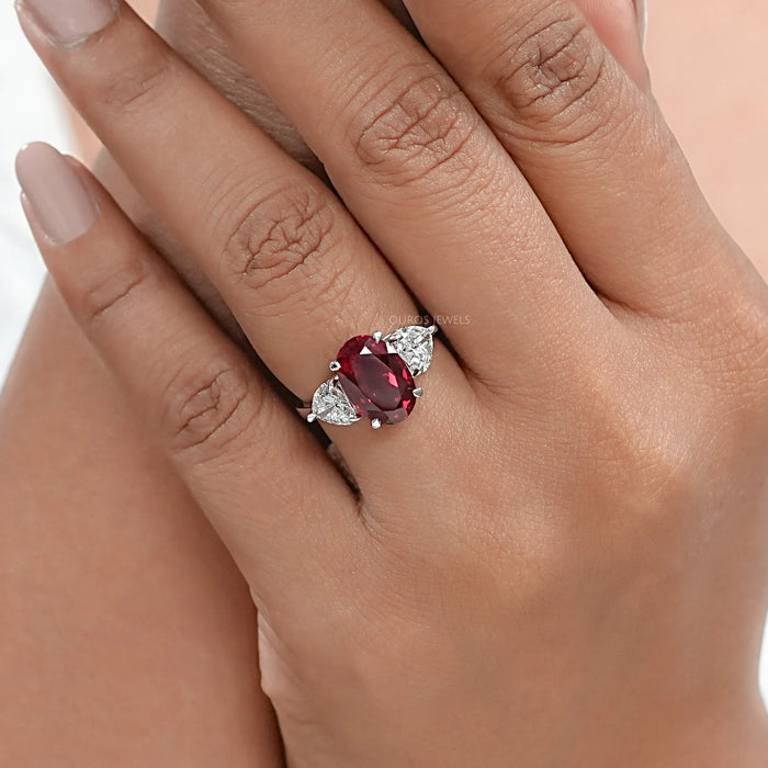 oval ruby gemstone with heart cut diamond 3 stone engagement ring on model's finger