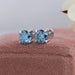 Blue Round Diamond Stud Earrings placed on a soft pink fabric background, displaying the brilliant blue diamonds and silver settings.