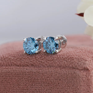 Blue Round Diamond Stud Earrings placed on a soft pink fabric background, displaying the brilliant blue diamonds and silver settings.