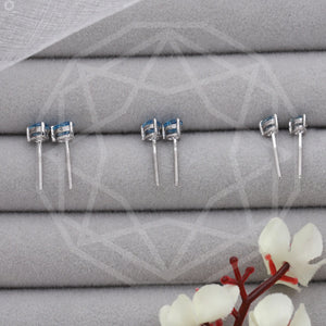 Blue Pear Shaped Diamond Stud Earrings displayed in a row, each earring set against a soft, gray fabric background with a delicate floral accent in the foreground.